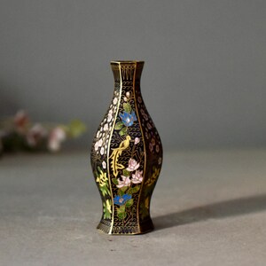 May include: A black decorative vase with a floral and bird design. The vase has a narrow neck and a flared base. The flowers are in shades of blue, pink, and white, with a yellow bird. The vase is approximately 6 inches tall.