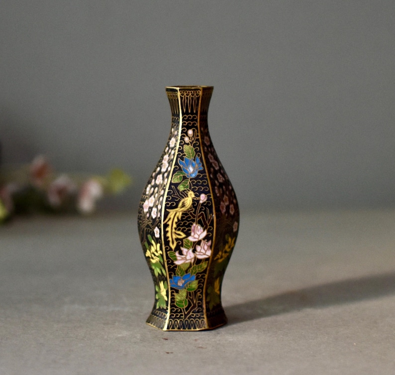 May include: A black decorative vase with a floral and bird design. The vase has a narrow neck and a flared base. The flowers are in shades of blue, pink, and white, with a yellow bird. The vase is approximately 6 inches tall.