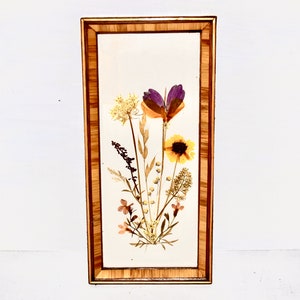 May include: A framed pressed flower art piece with a variety of dried flowers in shades of purple, yellow, and white. The flowers are arranged in a bouquet shape and are set against a white background. The frame is made of wood and has a natural finish.