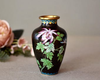May include: A small black cloisonné vase with a floral design. The vase is decorated with pink, white, and blue flowers and green leaves. The vase has a gold rim.