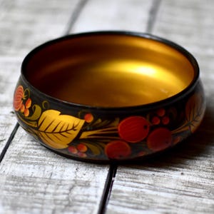 May include: A round wooden bowl with a black exterior and a smooth gold interior. The outer surface features a hand-painted design of golden leaves and clusters of red berries, creating a contrast against the dark background of the bowl.