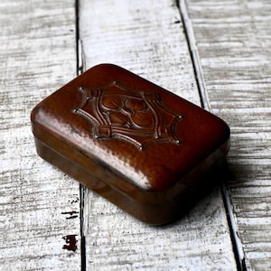 May include: A rectangular, brown wooden box with a hammered texture and a decorative floral design on the lid. The box is closed and sits on a weathered, white-painted wooden surface.
