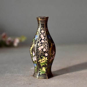 May include: A decorative black vase with a floral design. The vase features a slender neck and a flared base. The floral pattern includes pink, blue, and yellow flowers with gold accents. The vase is approximately 6 inches tall.