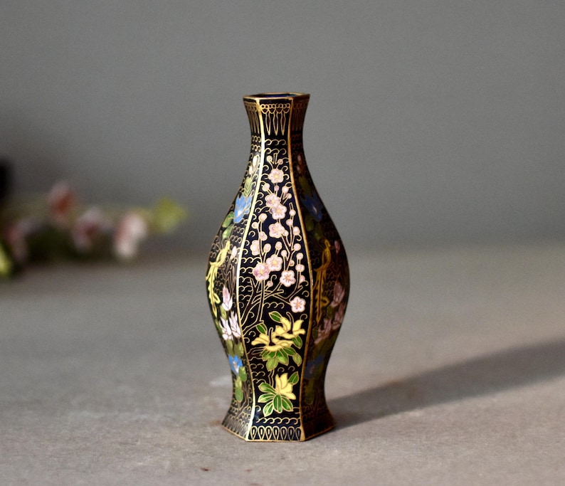 May include: A decorative black vase with a floral design. The vase features a slender neck and a flared base. The floral pattern includes pink, blue, and yellow flowers with gold accents. The vase is approximately 6 inches tall.