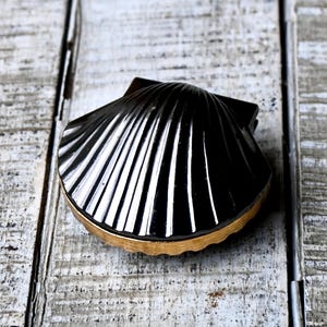 May include: A black seashell-shaped box with a gold-colored rim. The box has a glossy finish and is resting on a weathered, white-painted wooden surface. The shell design features prominent, vertical ridges.
