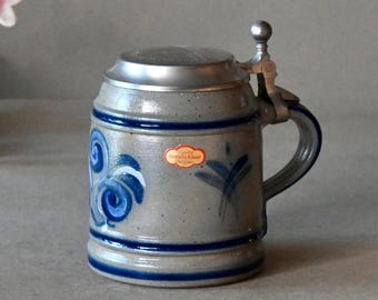May include: A stoneware beer stein with a hinged metal lid and handle. The mug is gray with blue painted floral designs and blue bands. A small red and gold sticker is on the front.