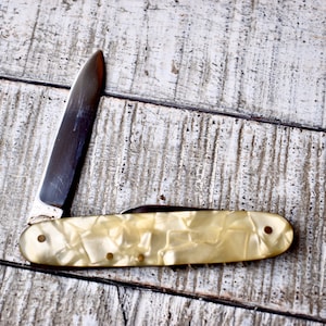 May include: A vintage pocket knife with a white pearl-like handle and a silver blade. The knife is closed and lying on a white wooden surface.