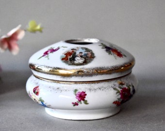May include: A white porcelain trinket box with a lid, featuring a gold rim and floral and classical scene designs. The box has a round base and a slightly domed lid with a central opening.