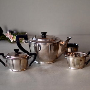 Może przedstawiać: Srebrny zestaw do herbaty z czarnymi uchwytami. Zestaw zawiera czajnik, dzbanek do śmietanki i cukiernicę.