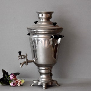 May include: A silver samovar with a black handle and a spout. The samovar is sitting on a gray surface with a bouquet of pink flowers in front of it.