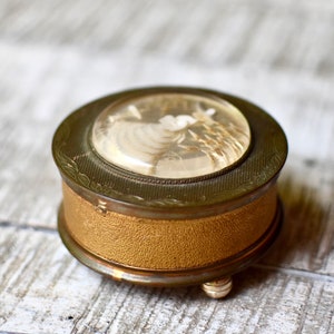 May include: A small, round, gold-colored music box with a clear lid. The lid features a white cameo of a woman with flowers. The music box has a textured, gold-colored base.