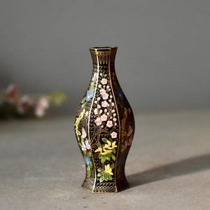 May include: A black decorative vase with a floral design. The vase features a slender neck and a flared base. The flowers are in shades of pink, blue, and yellow, with gold accents. The vase is approximately 6 inches tall.
