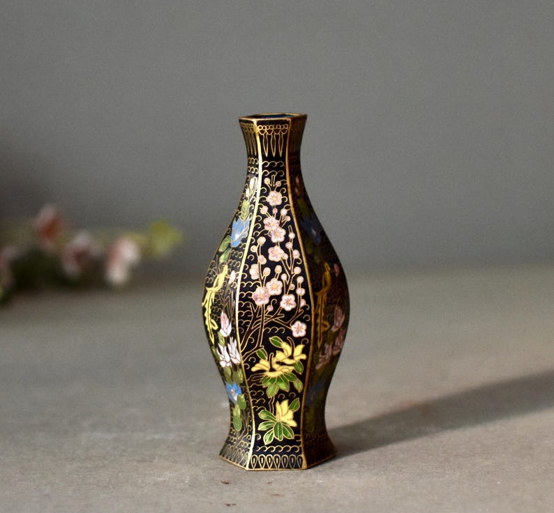 May include: A black decorative vase with a floral design. The vase features a slender neck and a flared base. The flowers are in shades of pink, blue, and yellow, with gold accents. The vase is approximately 6 inches tall.