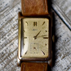 Op de afbeelding: Een vintage goudkleurig polshorloge met een rechthoekige wijzerplaat en een bruine leren band. De wijzerplaat van het horloge heeft gouden wijzers en markeringen, met het nummer II duidelijk weergegeven. Het horloge heeft een kleine secondenwijzer en de merknaam "Mara" is zichtbaar.