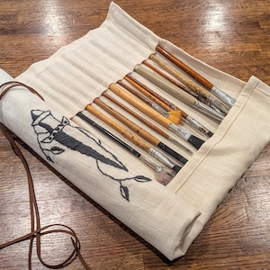 Puede incluir: Un estuche enrollado de lona beige con una correa de cuero marrón, que contiene varios pinceles. El estuche está decorado con una ilustración en blanco y negro de un pez. Los pinceles tienen mangos de madera y virolas metálicas.