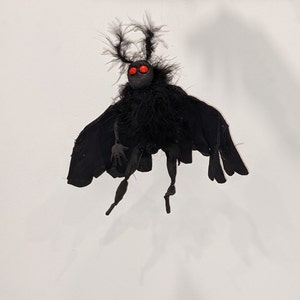 May include: A black moth puppet with red eyes and feathery wings. The puppet is suspended in mid-air against a white background.
