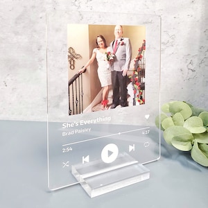 May include: A clear acrylic photo frame with a white background and a black and white photo of a couple. The frame has a music player design with the song title "She's Everything" by Brad Paisley and play, pause, and skip buttons.