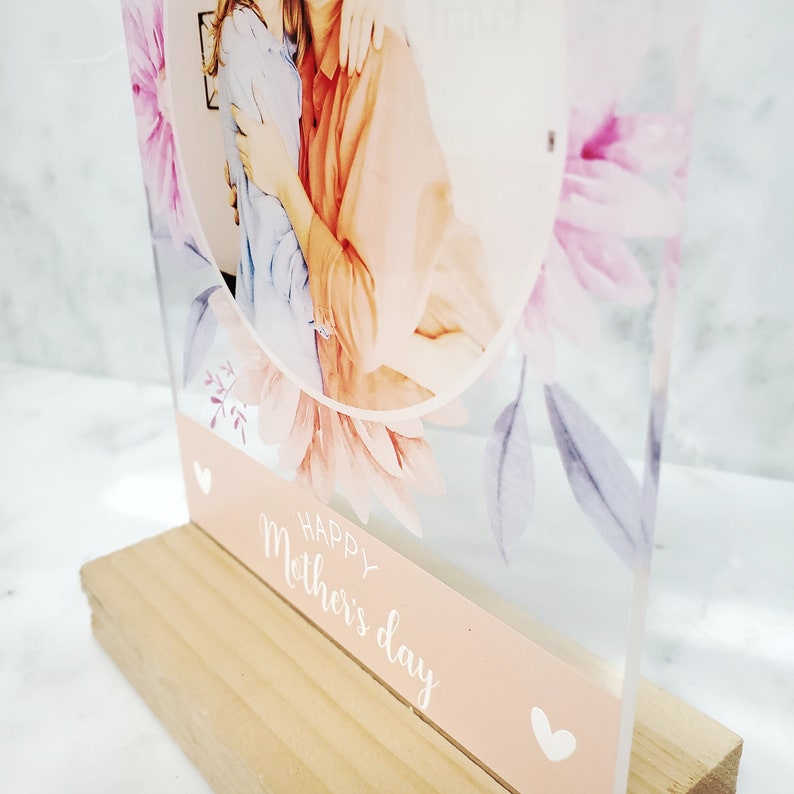 May include: A clear acrylic photo frame with a floral design and a photo of two people hugging. The frame has a pink and white floral design and the text "Happy Mother's Day" on the bottom.