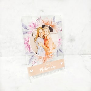 May include: A clear acrylic photo frame with a floral design and a photo of two women hugging. The frame has a pink and white background with the text "Happy Mother's Day" printed on it.