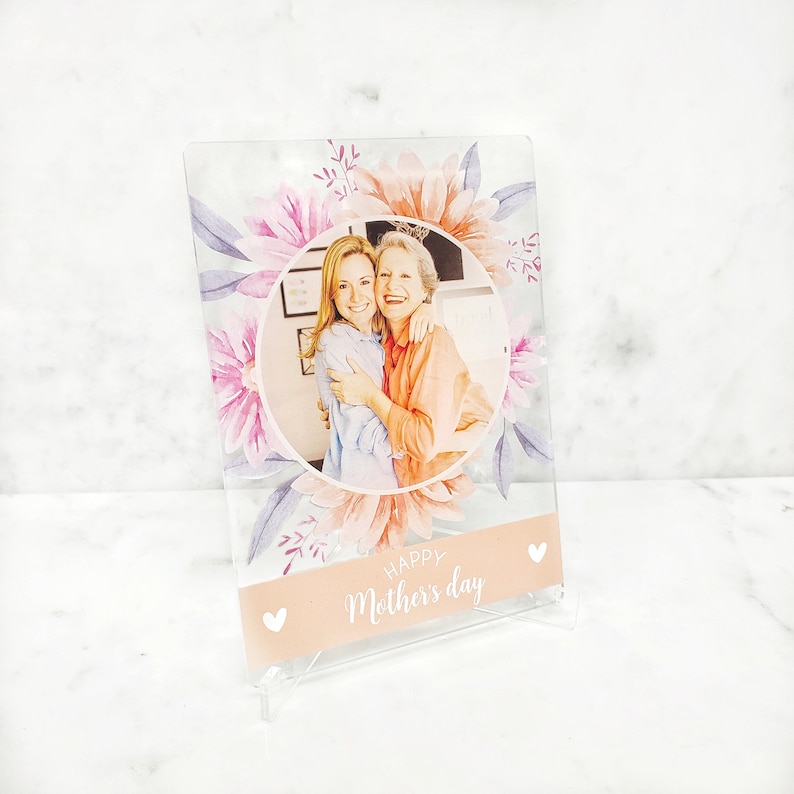 May include: A clear acrylic photo frame with a floral design and a photo of two women hugging. The frame has a pink and white background with the text "Happy Mother's Day" printed on it.
