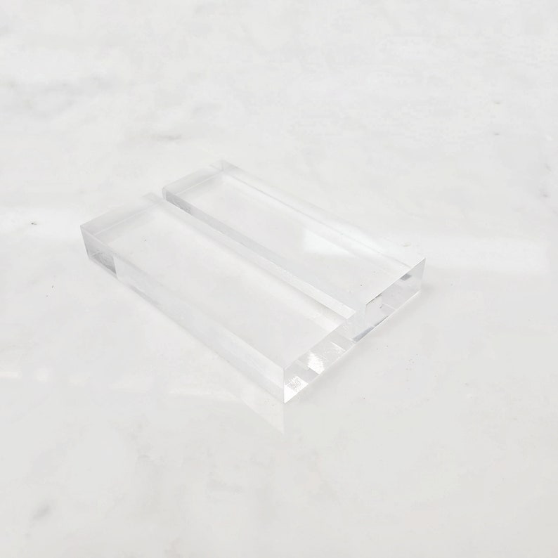 May include: A clear acrylic display stand with two slots for holding items, such as business cards or small photos.