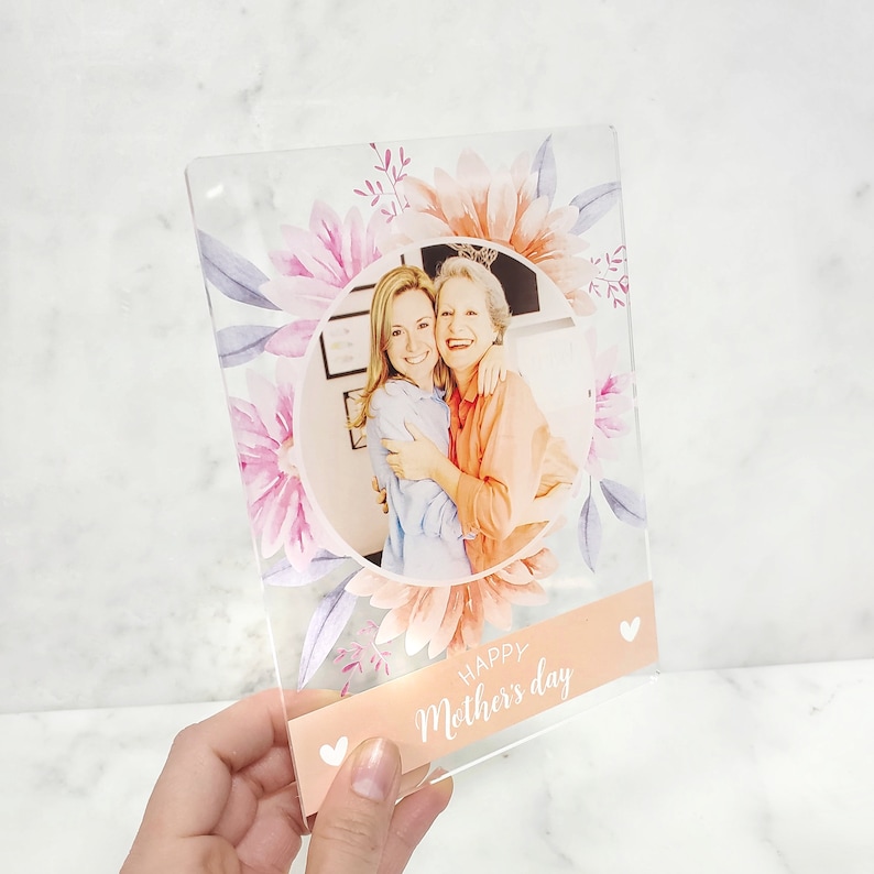 May include: A clear acrylic photo frame with a floral design and a photo of two women smiling. The frame has a pink and white floral design with a peach background. The text "Happy Mother's Day" is printed on the bottom of the frame.