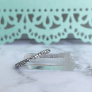 May include: A silver band with a row of small, clear cubic zirconia stones. The band is resting on a clear quartz crystal. The text "SIMPLYBRIDALL" is visible in the lower right corner of the image.