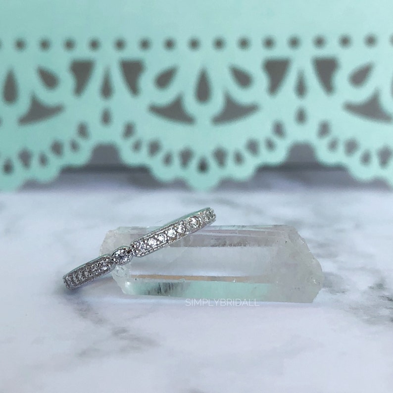 May include: A silver band with a row of small, clear cubic zirconia stones. The band is resting on a clear quartz crystal. The text "SIMPLYBRIDALL" is visible in the lower right corner of the image.