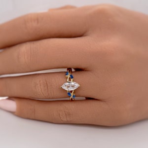 Puede incluir: Un anillo de oro con una piedra central de forma marquesa transparente y piedras azules más pequeñas en la banda.