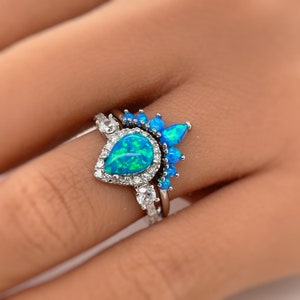 May include: A silver ring set with a teardrop-shaped blue opal surrounded by smaller clear stones. A second ring with a row of smaller blue opals sits above the first ring.