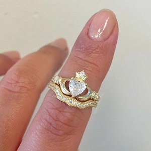 Può includere: Un anello d'oro con una pietra trasparente a forma di cuore e una corona in cima. L'anello è indossato su un dito con una fascia abbinata.