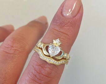 May include: A gold ring set with a heart-shaped clear stone and a crown on top. The ring is worn on a finger with a matching band.
