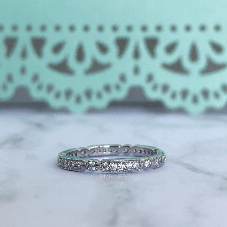 May include: A silver band ring with a row of small round and rectangular clear stones. The ring is on a white marble surface. "SIMPLYBRIDALL" is printed in black below the ring.