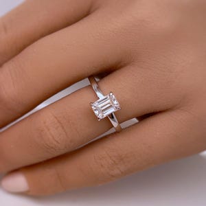 May include: A silver ring with a rectangular, emerald-cut diamond. The ring is set on a slender band and is displayed on a hand. The diamond is clear and reflects light.