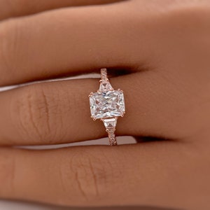 May include: A rose gold engagement ring with a large, rectangular, clear center stone and two smaller, triangular, clear stones on either side. The band is set with smaller, clear stones.