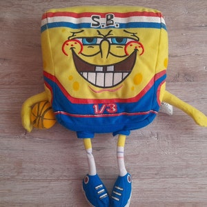 Puede incluir: Juguete de peluche amarillo de Bob Esponja con un jersey de baloncesto azul con el texto "S.B. 1/3". Está sosteniendo un balón de baloncesto y tiene una gran sonrisa en la cara.