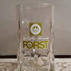 May include: Clear glass beer stein with a textured design and the text "FORST" in gold lettering. The stein has a square logo with a mountain design. The stein is approximately 6 inches tall.