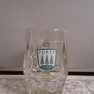 May include: Clear glass beer mug with a textured design. The mug features the word "FORST" above a green emblem with three stylized pine trees. The mug is designed for beer and other beverages.