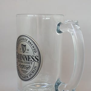 May include: Clear glass Guinness beer mug with a handle. The mug features a circular logo with the words "Guinness Extra Stout" and "Dublin" in black and white. The mug has a decorative base.