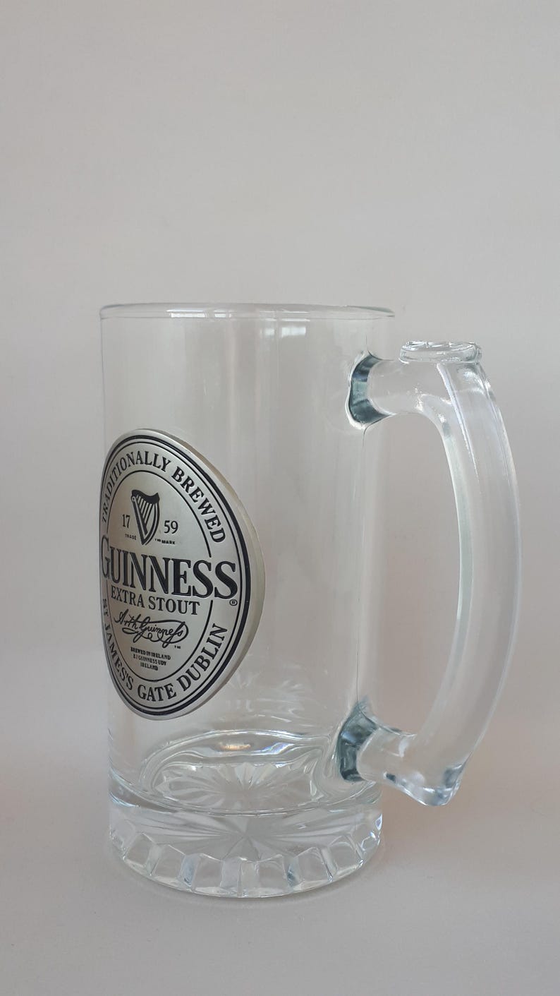 May include: Clear glass Guinness beer mug with a handle. The mug features a circular logo with the words "Guinness Extra Stout" and "Dublin" in black and white. The mug has a decorative base.