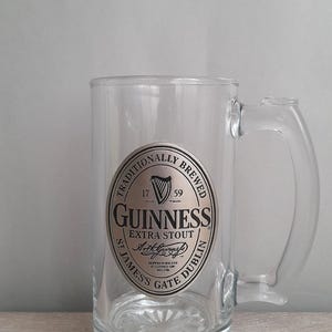 May include: A clear glass beer mug with a handle. The mug features a silver and black Guinness Extra Stout label with the text "Traditionally Brewed" and "St. James's Gate Dublin". The mug is empty and sits on a wooden surface.