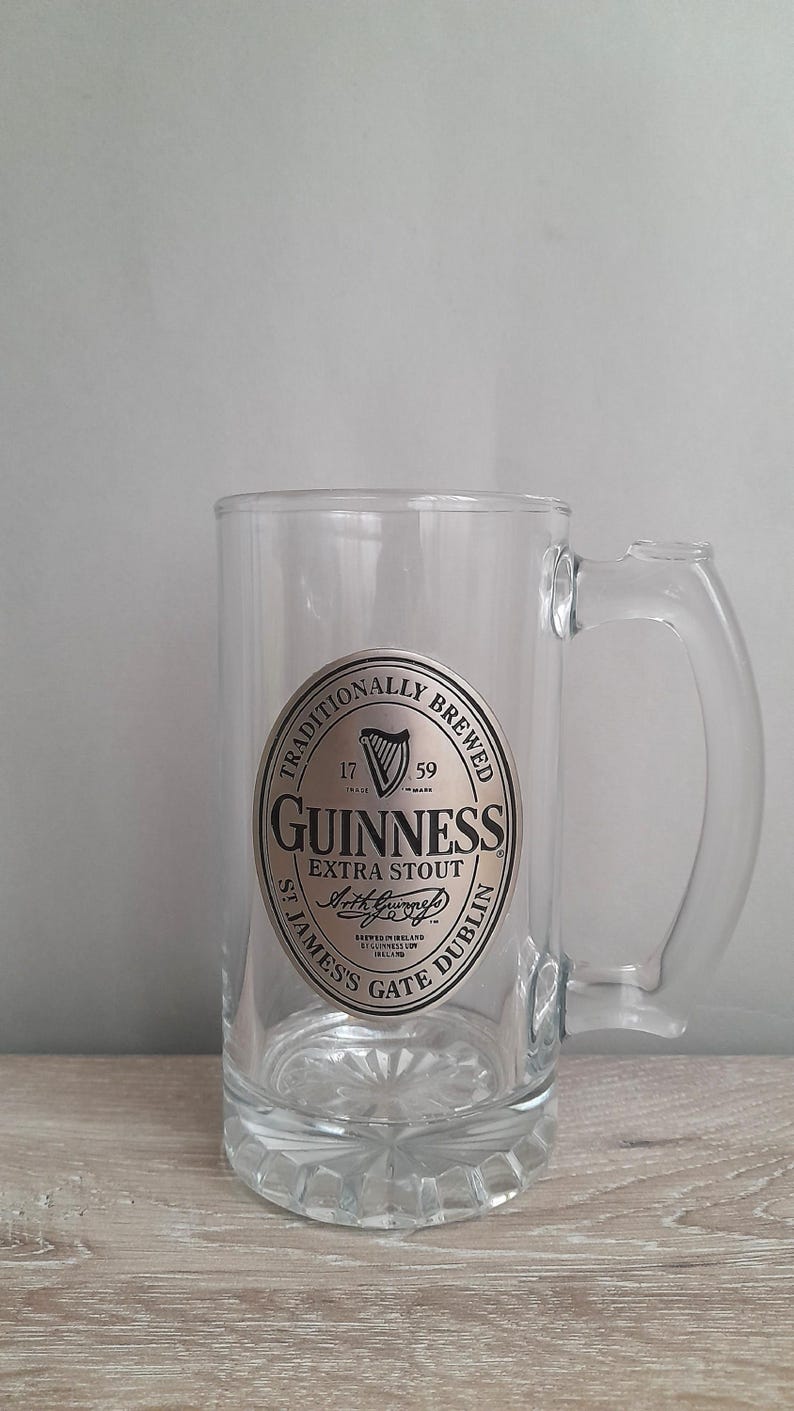 May include: A clear glass beer mug with a handle. The mug features a silver and black Guinness Extra Stout label with the text "Traditionally Brewed" and "St. James's Gate Dublin". The mug is empty and sits on a wooden surface.