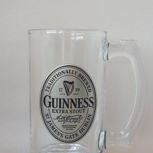 May include: Clear glass Guinness beer mug with a handle. The mug features the Guinness logo in a circular design with the words "Traditionally Brewed" and "Extra Stout". The mug is designed for beer.