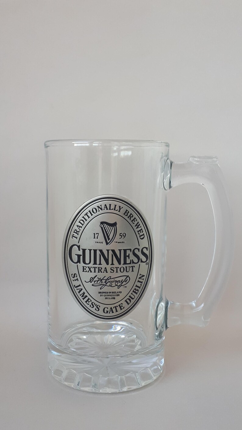May include: Clear glass Guinness beer mug with a handle. The mug features the Guinness logo in a circular design with the words "Traditionally Brewed" and "Extra Stout". The mug is designed for beer.