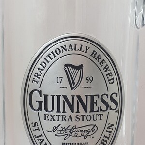 May include: Clear glass beer mug with a handle. The mug features a silver and black Guinness logo with the words "GUINNESS EXTRA STOUT" and "ST JAMES'S GATE DUBLIN" printed on it. The mug is empty.