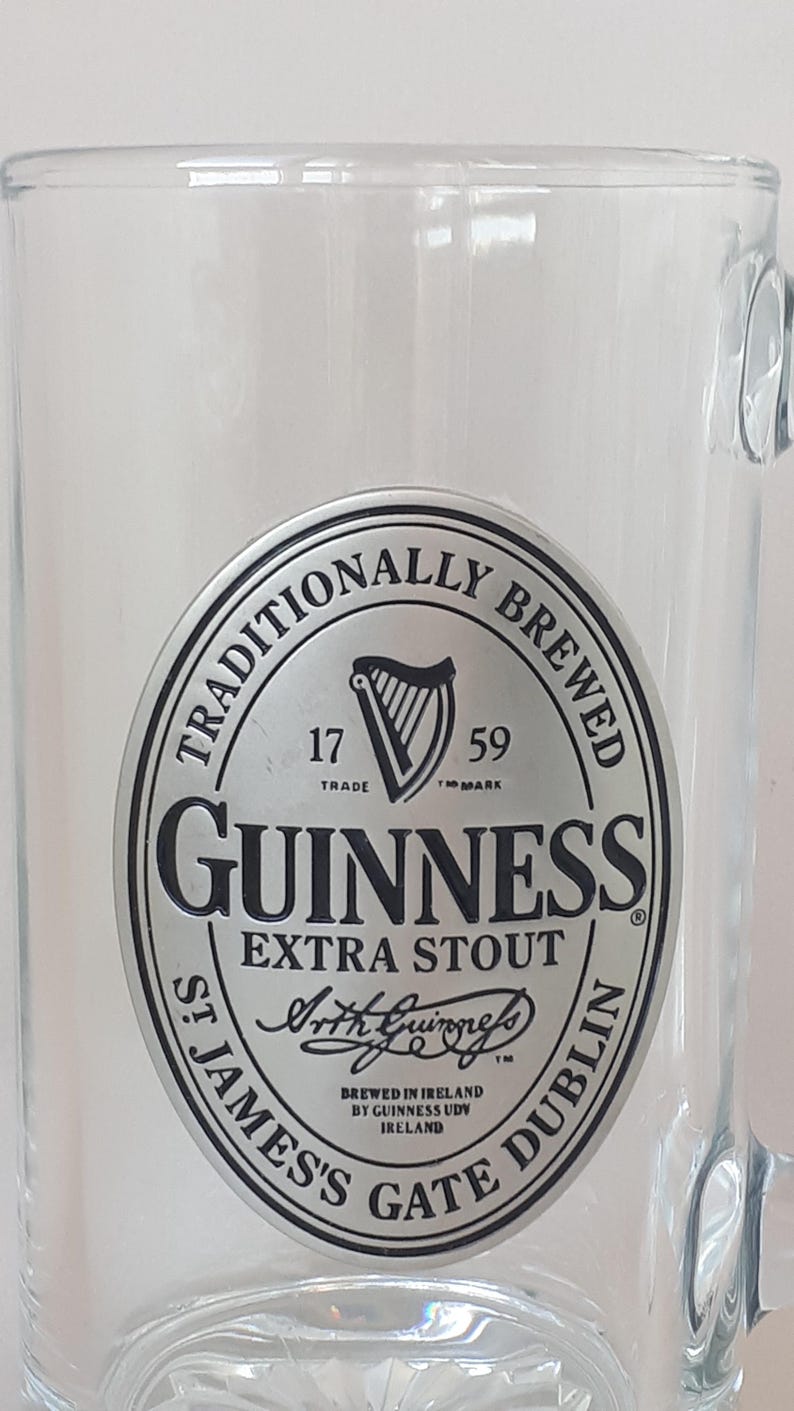 May include: Clear glass beer mug with a handle. The mug features a silver and black Guinness logo with the words "GUINNESS EXTRA STOUT" and "ST JAMES'S GATE DUBLIN" printed on it. The mug is empty.
