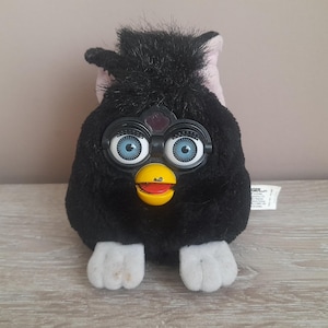 May include: A black Furby toy with large blue eyes, a yellow beak, and white feet. The toy has a tuft of black fur on top and a pinkish-purple area behind its head. A white tag is attached to the side.