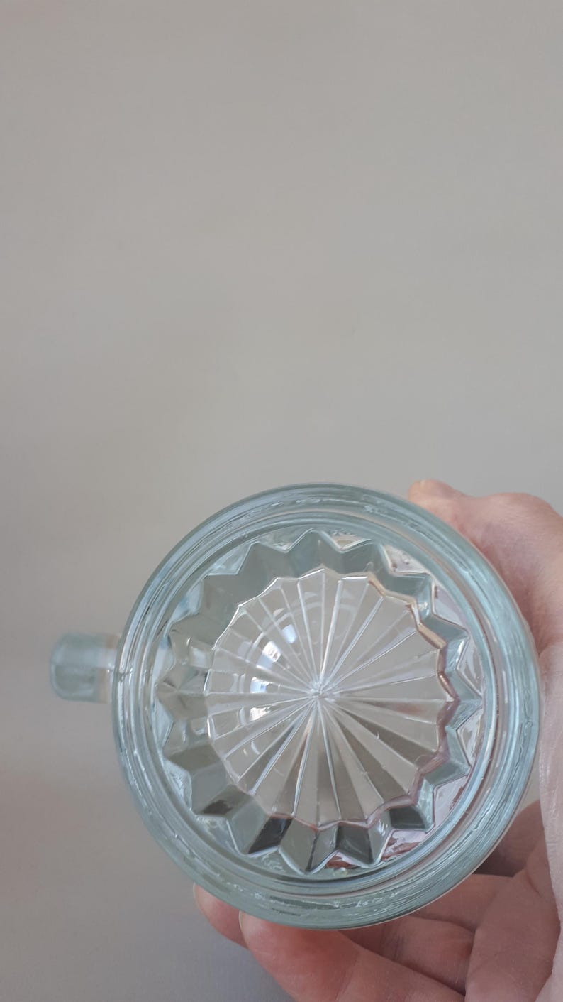 May include: Clear glass mug with a handle, featuring a starburst pattern on the base. The glass has a slightly bluish tint. The mug is designed with a rounded shape and a small handle for easy gripping.