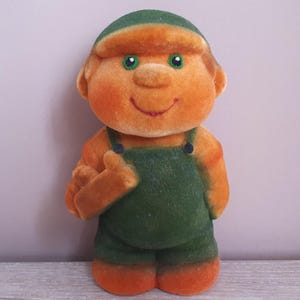 Op de afbeelding: Een vintage pluche pop met een oranje gezicht, handen en voeten, gekleed in een groene overall en pet. De pop heeft groene ogen en een glimlachende uitdrukking, met één hand omhoog in een duim omhoog gebaar.