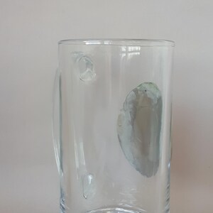 May include: A clear glass beer mug with a handle. The mug has a decorative, cut-glass base and a cylindrical body. There are a few small imperfections on the glass. The mug is empty and sits against a plain background.